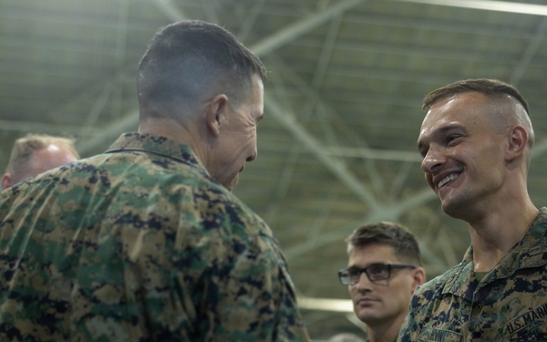 Two U.S. Marines in MARPAT camouflage uniforms are shaking hands in what appears to be a hangar or large indoor military facility. The Marine on the right is smiling broadly, suggesting a positive interaction such as a commendation, award, or greeting ceremony. Several other Marines are visible in the background observing the exchange.