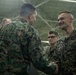 Two U.S. Marines in MARPAT camouflage uniforms are shaking hands in what appears to be a hangar or large indoor military facility. The Marine on the right is smiling broadly, suggesting a positive interaction such as a commendation, award, or greeting ceremony. Several other Marines are visible in the background observing the exchange.