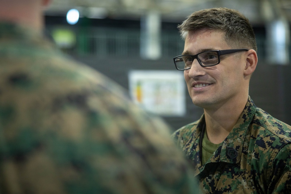 A male military service member wearing MARPAT (Marine pattern) digital camouflage uniform is engaged in conversation with another uniformed individual whose back is to the camera. The subject is smiling and wearing black-framed rectangular glasses, suggesting a relaxed, informal interaction. The setting appears to be an indoor military facility or gymnasium.