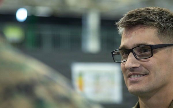 A male military service member wearing MARPAT (Marine pattern) digital camouflage uniform is engaged in conversation with another uniformed individual whose back is to the camera. The subject is smiling and wearing black-framed rectangular glasses, suggesting a relaxed, informal interaction. The setting appears to be an indoor military facility or gymnasium.