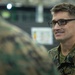A male military service member wearing MARPAT (Marine pattern) digital camouflage uniform is engaged in conversation with another uniformed individual whose back is to the camera. The subject is smiling and wearing black-framed rectangular glasses, suggesting a relaxed, informal interaction. The setting appears to be an indoor military facility or gymnasium.