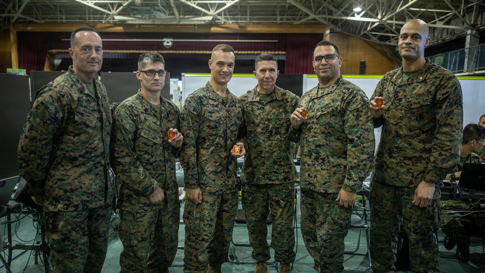 Six military personnel in U.S. Marine Corps MARPAT (digital camouflage) uniforms pose together for a group photo in what appears to be a large indoor facility or gymnasium. Several of the individuals are holding small red/orange challenge coins or medallions. The setting appears to be a military operations or command center environment with workstations visible in the background.