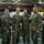 Six military personnel in U.S. Marine Corps MARPAT (digital camouflage) uniforms pose together for a group photo in what appears to be a large indoor facility or gymnasium. Several of the individuals are holding small red/orange challenge coins or medallions. The setting appears to be a military operations or command center environment with workstations visible in the background.