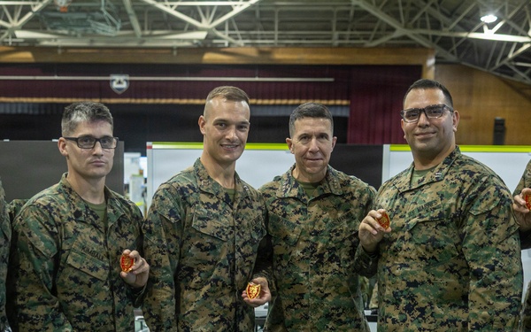 Six military personnel in U.S. Marine Corps MARPAT (digital camouflage) uniforms pose together for a group photo in what appears to be a large indoor facility or gymnasium. Several of the individuals are holding small red/orange challenge coins or medallions. The setting appears to be a military operations or command center environment with workstations visible in the background.