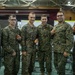 Six military personnel in U.S. Marine Corps MARPAT (digital camouflage) uniforms pose together for a group photo in what appears to be a large indoor facility or gymnasium. Several of the individuals are holding small red/orange challenge coins or medallions. The setting appears to be a military operations or command center environment with workstations visible in the background.