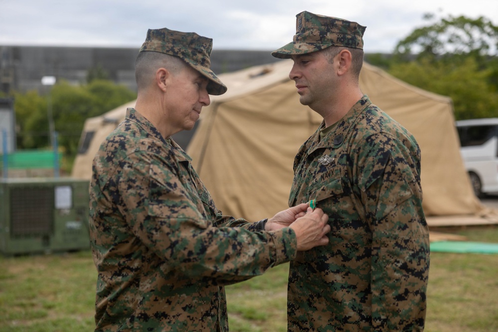 Two military personnel in MARPAT woodland camouflage uniforms are engaged in a medal or award pinning ceremony outdoors. The individual on the left is pinning a decoration onto the chest of the individual on the right, who is identified by a 'NAVY' tape on his uniform. The setting appears to be a field environment with military tents and equipment visible in the background.