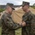 Two military personnel in MARPAT woodland camouflage uniforms are engaged in a medal or award pinning ceremony outdoors. The individual on the left is pinning a decoration onto the chest of the individual on the right, who is identified by a 'NAVY' tape on his uniform. The setting appears to be a field environment with military tents and equipment visible in the background.