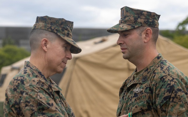 Two military personnel in MARPAT woodland camouflage uniforms are engaged in a medal or award pinning ceremony outdoors. The individual on the left is pinning a decoration onto the chest of the individual on the right, who is identified by a 'NAVY' tape on his uniform. The setting appears to be a field environment with military tents and equipment visible in the background.