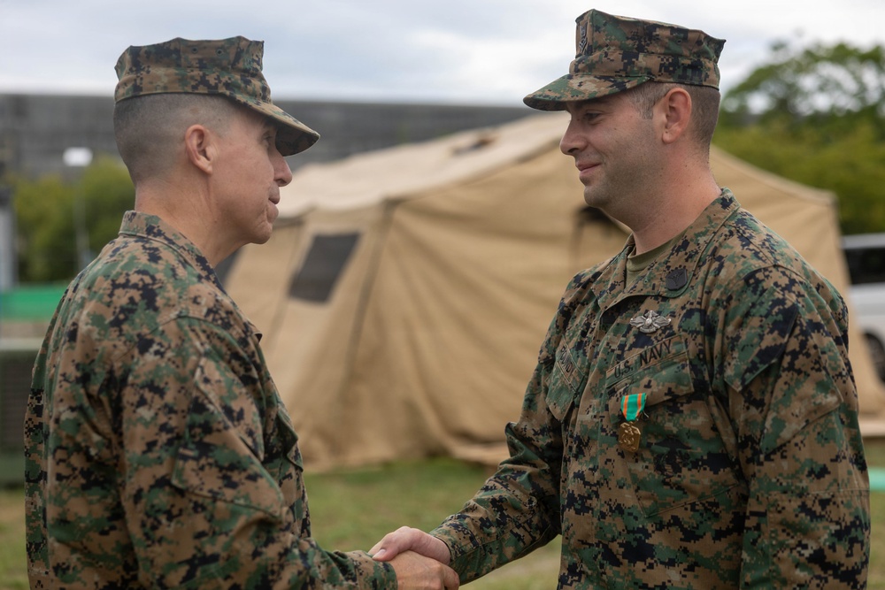 Two military personnel in MARPAT (Marine Pattern) camouflage uniforms are shaking hands in what appears to be an award or recognition ceremony. The individual on the right is wearing a U.S. Navy uniform with a medal pinned to his chest, suggesting he has just received a commendation. The setting appears to be an outdoor military field environment with a tan military tent visible in the background.