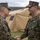 Two military personnel in MARPAT (Marine Pattern) camouflage uniforms are shaking hands in what appears to be an award or recognition ceremony. The individual on the right is wearing a U.S. Navy uniform with a medal pinned to his chest, suggesting he has just received a commendation. The setting appears to be an outdoor military field environment with a tan military tent visible in the background.