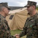 Two military personnel in MARPAT (Marine Pattern) camouflage uniforms are shaking hands in what appears to be an award or recognition ceremony. The individual on the right is wearing a U.S. Navy uniform with a medal pinned to his chest, suggesting he has just received a commendation. The setting appears to be an outdoor military field environment with a tan military tent visible in the background.