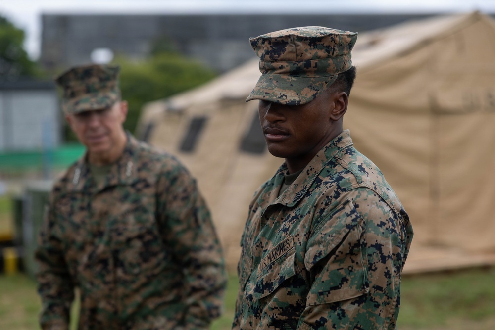 Two U.S. Marines in MARPAT woodland camouflage uniforms are standing outdoors near military field tents. The foreground Marine is in sharp focus while a second Marine stands in the background to the left. The setting appears to be a temporary field operations area or training exercise site.