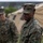 Two U.S. Marines in MARPAT woodland camouflage uniforms are standing outdoors near military field tents. The foreground Marine is in sharp focus while a second Marine stands in the background to the left. The setting appears to be a temporary field operations area or training exercise site.
