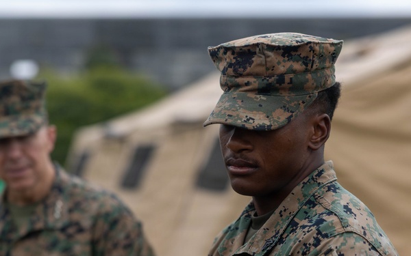 Two U.S. Marines in MARPAT woodland camouflage uniforms are standing outdoors near military field tents. The foreground Marine is in sharp focus while a second Marine stands in the background to the left. The setting appears to be a temporary field operations area or training exercise site.
