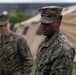 Two U.S. Marines in MARPAT woodland camouflage uniforms are standing outdoors near military field tents. The foreground Marine is in sharp focus while a second Marine stands in the background to the left. The setting appears to be a temporary field operations area or training exercise site.