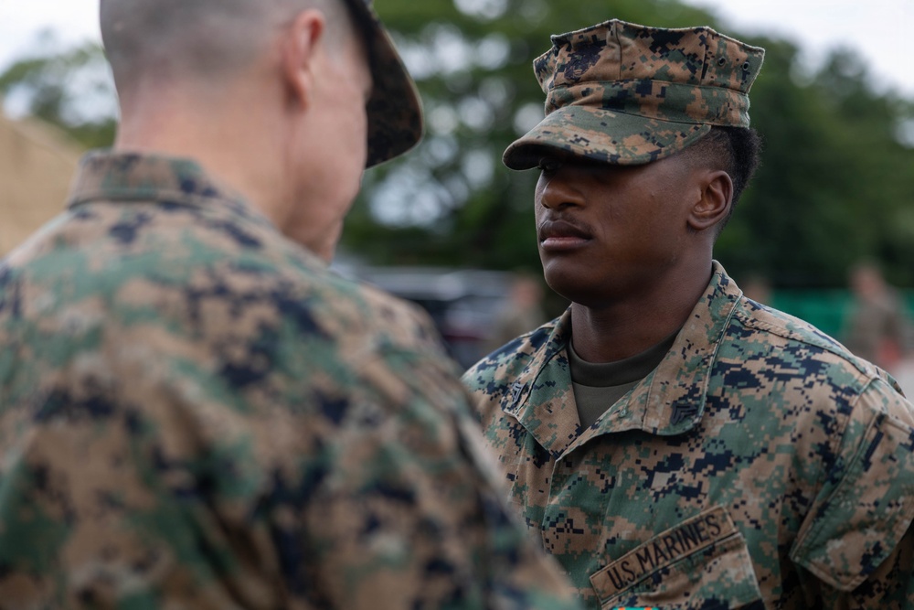 Two U.S. Marines in MARPAT (Marine Pattern) digital camouflage uniforms are engaged in a face-to-face interaction, likely a briefing or inspection. The Marine facing the camera wears a 'U.S. MARINES' tape on his uniform and a cover (cap) with the same camouflage pattern. The setting appears to be an outdoor military environment with trees and other personnel visible in the background.