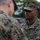 Two U.S. Marines in MARPAT (Marine Pattern) digital camouflage uniforms are engaged in a face-to-face interaction, likely a briefing or inspection. The Marine facing the camera wears a 'U.S. MARINES' tape on his uniform and a cover (cap) with the same camouflage pattern. The setting appears to be an outdoor military environment with trees and other personnel visible in the background.