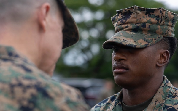 Two U.S. Marines in MARPAT (Marine Pattern) digital camouflage uniforms are engaged in a face-to-face interaction, likely a briefing or inspection. The Marine facing the camera wears a 'U.S. MARINES' tape on his uniform and a cover (cap) with the same camouflage pattern. The setting appears to be an outdoor military environment with trees and other personnel visible in the background.