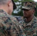 Two U.S. Marines in MARPAT (Marine Pattern) digital camouflage uniforms are engaged in a face-to-face interaction, likely a briefing or inspection. The Marine facing the camera wears a 'U.S. MARINES' tape on his uniform and a cover (cap) with the same camouflage pattern. The setting appears to be an outdoor military environment with trees and other personnel visible in the background.