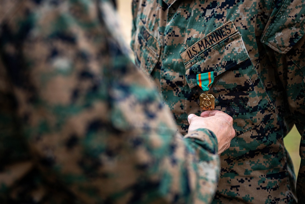 A U.S. Marines service member is being awarded a military medal during a formal pinning ceremony. A second person's hand is visible holding or pinning a gold medal with a teal and orange ribbon onto the recipient's MARPAT (Marine Pattern) camouflage uniform. The 'U.S. MARINES' tape is clearly visible on the uniform chest.