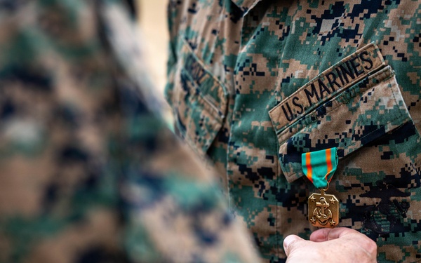 A U.S. Marines service member is being awarded a military medal during a formal pinning ceremony. A second person's hand is visible holding or pinning a gold medal with a teal and orange ribbon onto the recipient's MARPAT (Marine Pattern) camouflage uniform. The 'U.S. MARINES' tape is clearly visible on the uniform chest.