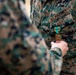 A U.S. Marines service member is being awarded a military medal during a formal pinning ceremony. A second person's hand is visible holding or pinning a gold medal with a teal and orange ribbon onto the recipient's MARPAT (Marine Pattern) camouflage uniform. The 'U.S. MARINES' tape is clearly visible on the uniform chest.