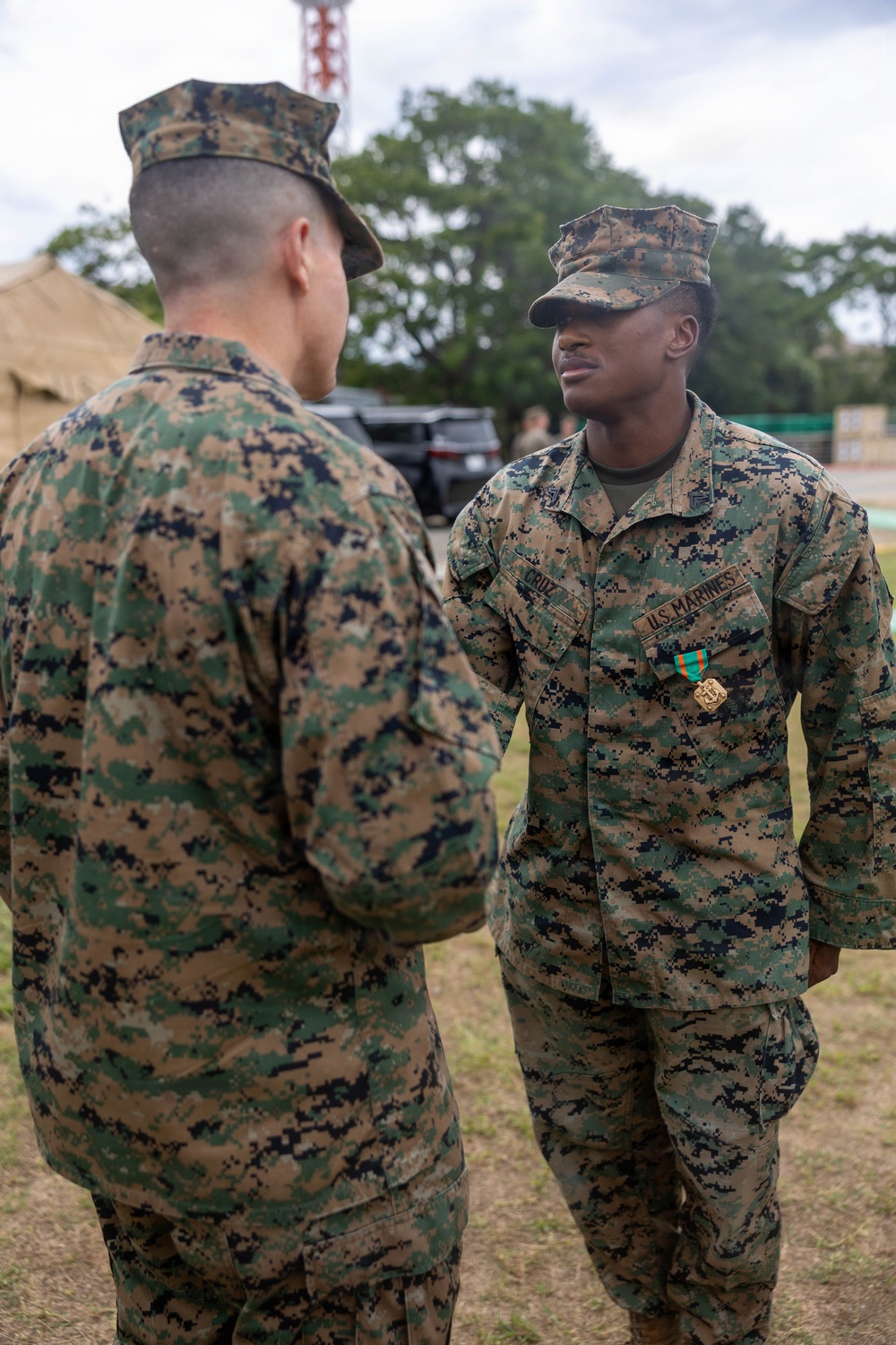 Two U.S. Marines in MARPAT (Marine Pattern) digital camouflage uniforms are engaged in a face-to-face interaction outdoors. The Marine facing the camera, identified by the name tape 'CRUZ' and a 'U.S. MARINES' tape, is wearing a medal with a green and teal ribbon, suggesting a recent award ceremony. The setting appears to be a field or base environment with tents, trees, and a communications tower visible in the background.