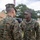 Two U.S. Marines in MARPAT (Marine Pattern) digital camouflage uniforms are engaged in a face-to-face interaction outdoors. The Marine facing the camera, identified by the name tape 'CRUZ' and a 'U.S. MARINES' tape, is wearing a medal with a green and teal ribbon, suggesting a recent award ceremony. The setting appears to be a field or base environment with tents, trees, and a communications tower visible in the background.