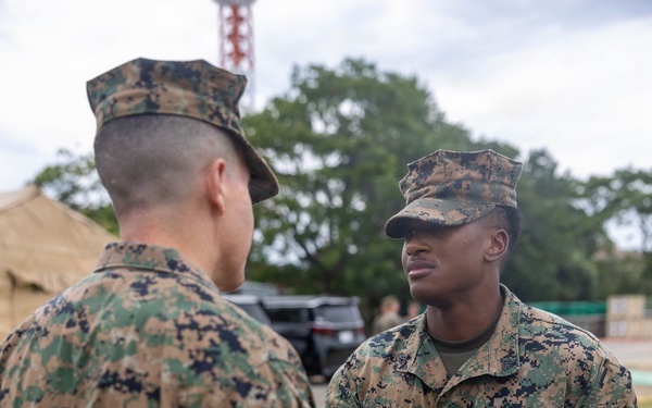 Two U.S. Marines in MARPAT (Marine Pattern) digital camouflage uniforms are engaged in a face-to-face interaction outdoors. The Marine facing the camera, identified by the name tape 'CRUZ' and a 'U.S. MARINES' tape, is wearing a medal with a green and teal ribbon, suggesting a recent award ceremony. The setting appears to be a field or base environment with tents, trees, and a communications tower visible in the background.