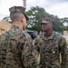 Two U.S. Marines in MARPAT (Marine Pattern) digital camouflage uniforms are engaged in a face-to-face interaction outdoors. The Marine facing the camera, identified by the name tape 'CRUZ' and a 'U.S. MARINES' tape, is wearing a medal with a green and teal ribbon, suggesting a recent award ceremony. The setting appears to be a field or base environment with tents, trees, and a communications tower visible in the background.