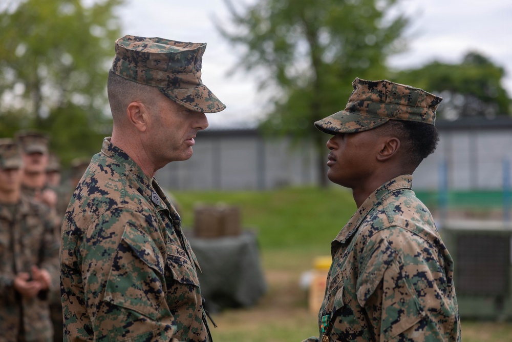 Two U.S. Marines in MARPAT digital camouflage uniforms face each other in what appears to be a formal military ceremony. The senior Marine on the left appears to be addressing or presenting something to the junior Marine on the right, who has a medal visible on their uniform. Additional Marines in uniform are visible in the blurred background.