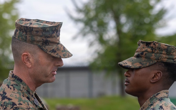 Two U.S. Marines in MARPAT digital camouflage uniforms face each other in what appears to be a formal military ceremony. The senior Marine on the left appears to be addressing or presenting something to the junior Marine on the right, who has a medal visible on their uniform. Additional Marines in uniform are visible in the blurred background.