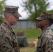 Two U.S. Marines in MARPAT digital camouflage uniforms face each other in what appears to be a formal military ceremony. The senior Marine on the left appears to be addressing or presenting something to the junior Marine on the right, who has a medal visible on their uniform. Additional Marines in uniform are visible in the blurred background.