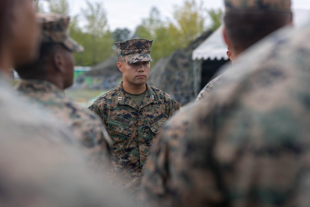 A U.S. Marine officer with the name tape 'JOHNSON' and visible captain's rank insignia is standing in the center of a group of fellow Marines in MARPAT woodland digital camouflage uniforms. The setting appears to be an outdoor field environment with camouflage netting and tents visible in the background. The officer appears to be addressing or briefing the surrounding Marines.