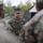 A U.S. Marine officer with the name tape 'JOHNSON' and visible captain's rank insignia is standing in the center of a group of fellow Marines in MARPAT woodland digital camouflage uniforms. The setting appears to be an outdoor field environment with camouflage netting and tents visible in the background. The officer appears to be addressing or briefing the surrounding Marines.