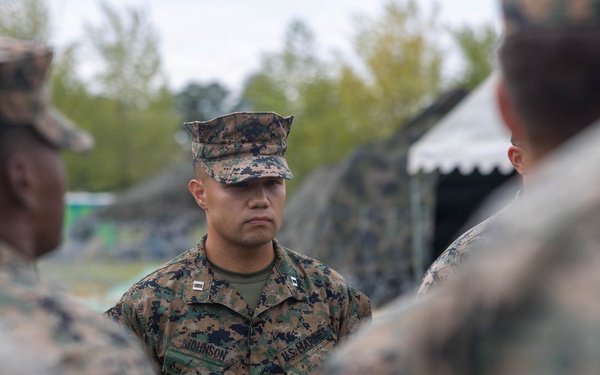 A U.S. Marine officer with the name tape 'JOHNSON' and visible captain's rank insignia is standing in the center of a group of fellow Marines in MARPAT woodland digital camouflage uniforms. The setting appears to be an outdoor field environment with camouflage netting and tents visible in the background. The officer appears to be addressing or briefing the surrounding Marines.
