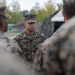 A U.S. Marine officer with the name tape 'JOHNSON' and visible captain's rank insignia is standing in the center of a group of fellow Marines in MARPAT woodland digital camouflage uniforms. The setting appears to be an outdoor field environment with camouflage netting and tents visible in the background. The officer appears to be addressing or briefing the surrounding Marines.