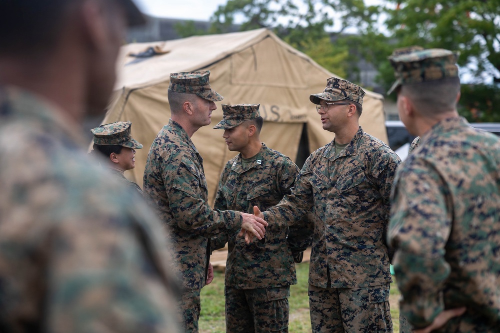 A group of U.S. Marines in MARPAT woodland camouflage uniforms are gathered outdoors near a tan military field tent marked 'U.S.' Two Marines in the center are shaking hands, suggesting a formal greeting, recognition, or introduction ceremony. The setting appears to be a military installation or field exercise area.