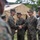 A group of U.S. Marines in MARPAT woodland camouflage uniforms are gathered outdoors near a tan military field tent marked 'U.S.' Two Marines in the center are shaking hands, suggesting a formal greeting, recognition, or introduction ceremony. The setting appears to be a military installation or field exercise area.
