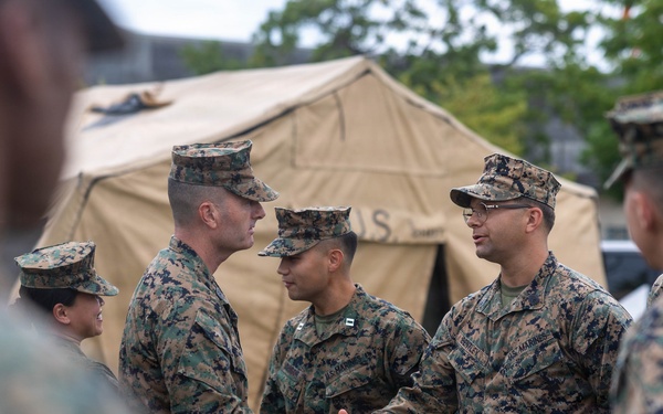 A group of U.S. Marines in MARPAT woodland camouflage uniforms are gathered outdoors near a tan military field tent marked 'U.S.' Two Marines in the center are shaking hands, suggesting a formal greeting, recognition, or introduction ceremony. The setting appears to be a military installation or field exercise area.