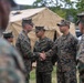 A group of U.S. Marines in MARPAT woodland camouflage uniforms are gathered outdoors near a tan military field tent marked 'U.S.' Two Marines in the center are shaking hands, suggesting a formal greeting, recognition, or introduction ceremony. The setting appears to be a military installation or field exercise area.