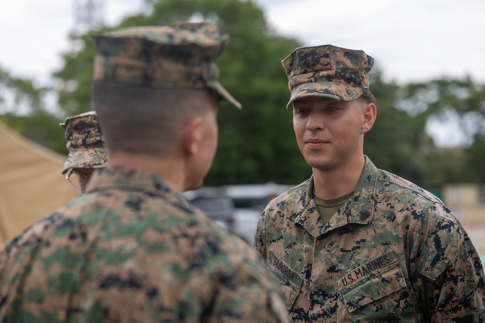 Two U.S. Marines in MARPAT (Marine Pattern) digital camouflage uniforms are engaged in a face-to-face conversation. The Marine facing the camera has a name tape reading 'TABORDA' and a 'U.S. MARINES' branch tape visible on his uniform. The setting appears to be an outdoor military area with tents and vehicles in the background.