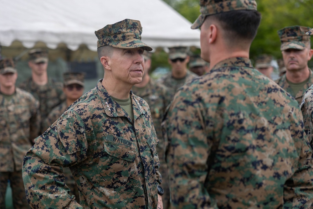 A group of U.S. Marines in MARPAT woodland digital camouflage uniforms are gathered outdoors near a white tent structure. The central figure, identified by a name tape reading 'WATSON,' appears to be a senior officer with two stars visible on the collar, indicating the rank of Major General. He is engaged in a face-to-face conversation with another Marine whose back is to the camera.