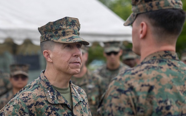 A group of U.S. Marines in MARPAT woodland digital camouflage uniforms are gathered outdoors near a white tent structure. The central figure, identified by a name tape reading 'WATSON,' appears to be a senior officer with two stars visible on the collar, indicating the rank of Major General. He is engaged in a face-to-face conversation with another Marine whose back is to the camera.