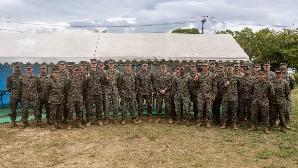 A large group of military personnel, approximately 25-30 individuals, pose for a formal group photograph in front of a large white tent structure. All personnel are wearing matching woodland camouflage uniforms and tan/brown boots, consistent with U.S. Marine Corps MARPAT pattern. Several individuals in the group appear to be holding small objects, possibly coins or medals, suggesting a recognition or award ceremony.