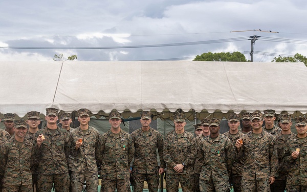 A large group of military personnel, approximately 25-30 individuals, pose for a formal group photograph in front of a large white tent structure. All personnel are wearing matching woodland camouflage uniforms and tan/brown boots, consistent with U.S. Marine Corps MARPAT pattern. Several individuals in the group appear to be holding small objects, possibly coins or medals, suggesting a recognition or award ceremony.