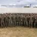 A large group of military personnel, approximately 25-30 individuals, pose for a formal group photograph in front of a large white tent structure. All personnel are wearing matching woodland camouflage uniforms and tan/brown boots, consistent with U.S. Marine Corps MARPAT pattern. Several individuals in the group appear to be holding small objects, possibly coins or medals, suggesting a recognition or award ceremony.