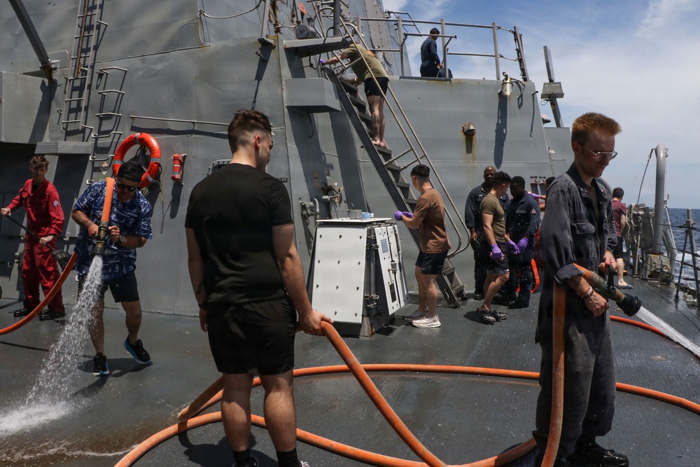 Naval personnel are conducting a washdown or cleaning operation on the deck of a gray military warship at sea. Multiple sailors in varied uniforms and civilian-style clothing are handling orange fire hoses, with water actively spraying across the deck. Additional crew members are visible on a stairway and in the background performing related tasks.