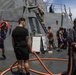Naval personnel are conducting a washdown or cleaning operation on the deck of a gray military warship at sea. Multiple sailors in varied uniforms and civilian-style clothing are handling orange fire hoses, with water actively spraying across the deck. Additional crew members are visible on a stairway and in the background performing related tasks.