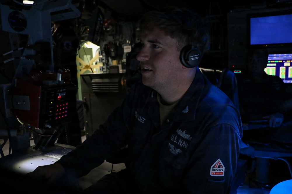 A U.S. Navy sailor is seated at a control station in a dimly lit operations room, wearing Sennheiser headphones and a navy blue uniform with 'U.S. NAVY' and a name tag reading 'BWANZ' visible. The environment appears to be a combat information center or bridge aboard a naval vessel, with multiple electronic displays and control panels visible. The sailor appears to be actively communicating or monitoring systems.